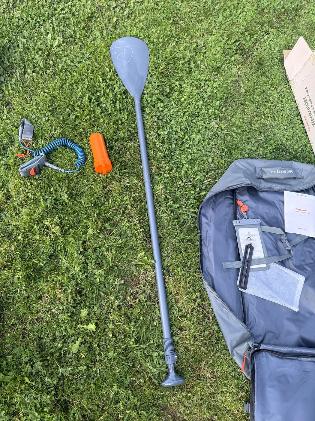 Assembled Retrospec paddle on the grass beside the coiled leash, orange repair kit canister and the open backpack with phone case